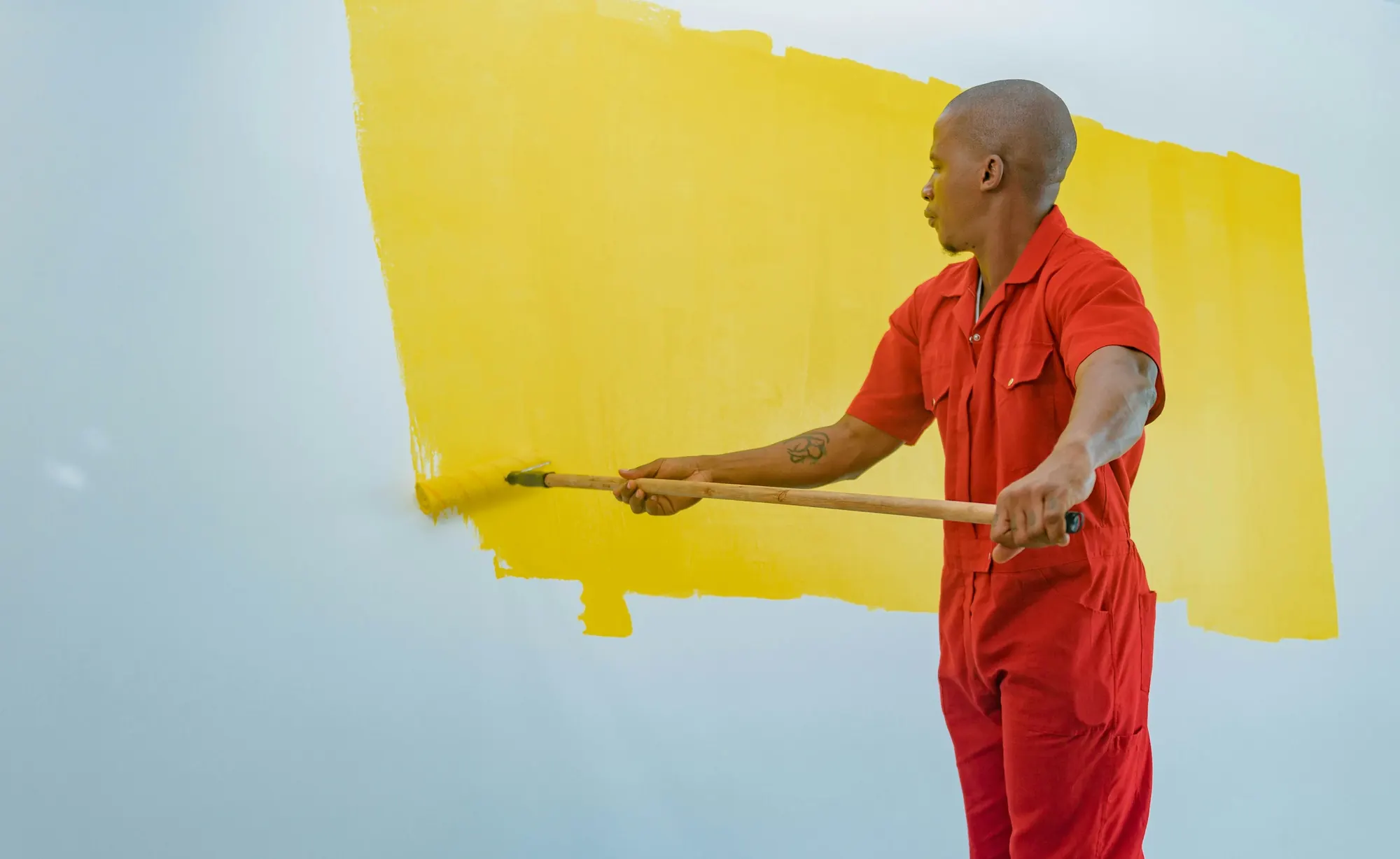 A painter in red coveralls rolling bright yellow paint onto a pale blue wall