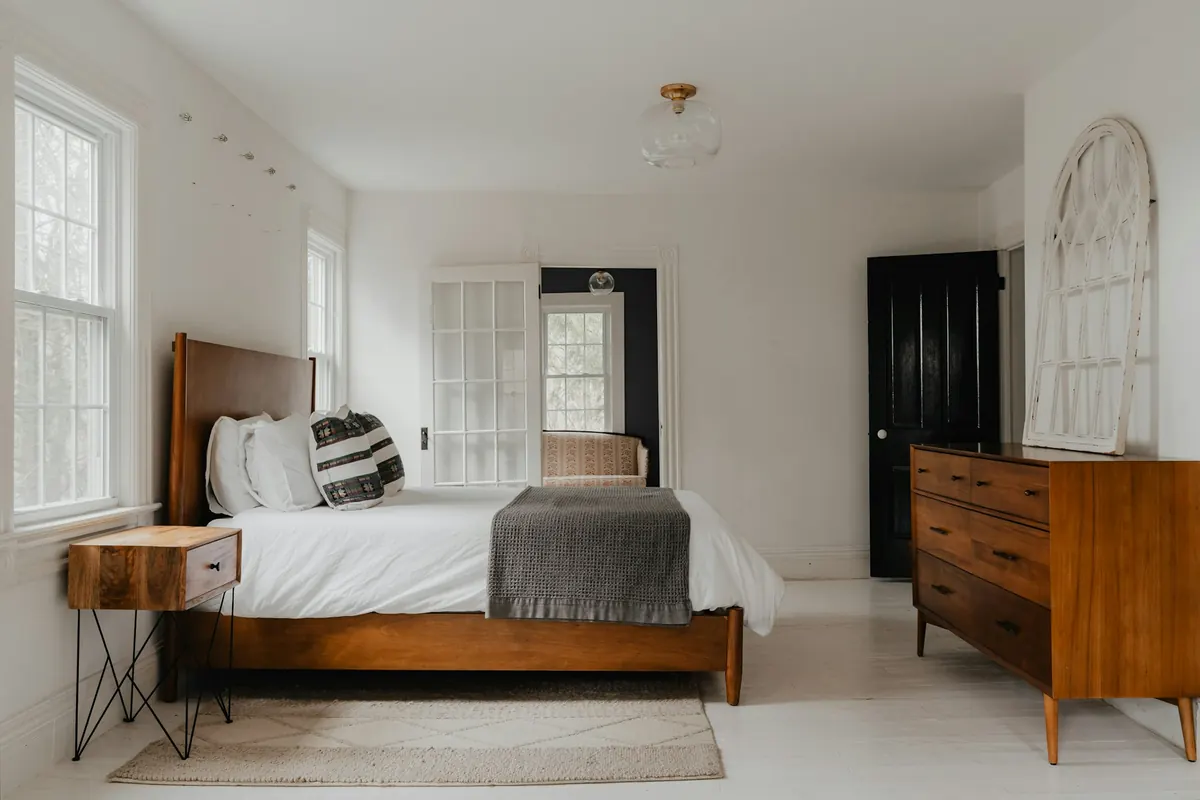 A bright white bedroom with a neatly made walnut bed, white linens and tall windows