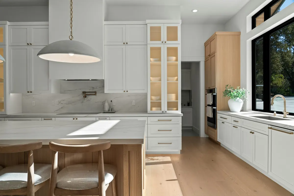 A spotless white kitchen with marble counters, a light-oak island and sunlight across the floor