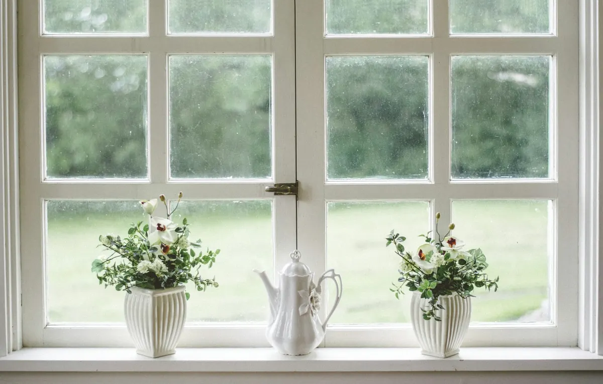 A white-framed window with vases of flowers on the sill and a green garden beyond