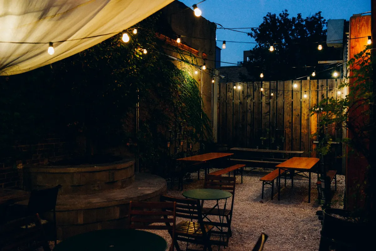 A courtyard patio at dusk with string lights, wooden tables and ivy-covered walls