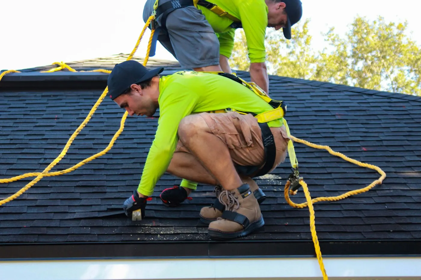 Two roofers in safety harnesses and high-visibility shirts working on a shingle roof