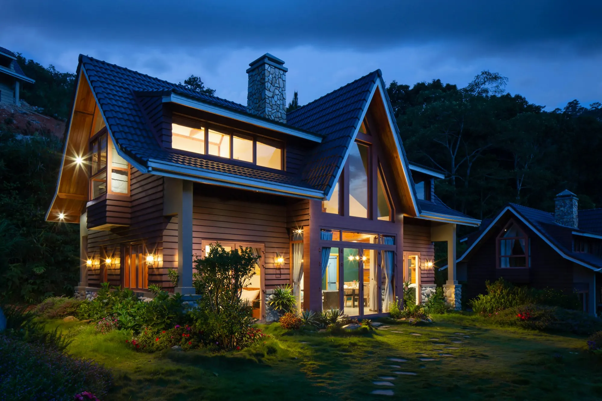 A wood-sided family home at dusk with warm lit windows and a dark shingle roof